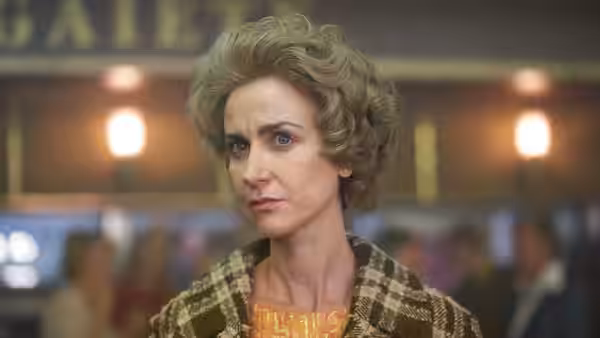 A curly-haired woman in a patterned coat looks to the side inside a brightly lit building
