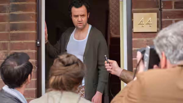 A man in a vest stands in his doorway as reporters hold microphones toward him