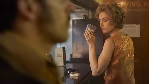 A woman in a gold dress raises a glass at a counter while glancing over her shoulder