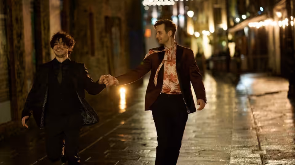 Two men walk hand in hand along a wet, lantern-lit street