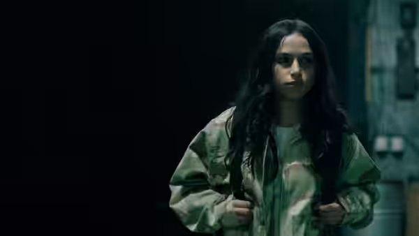 A young woman in a camouflage jacket stands in a dim corridor, gripping her backpack straps