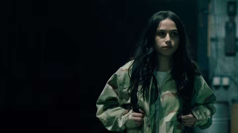 A young woman in a camouflage jacket stands in a dim corridor, gripping her backpack straps