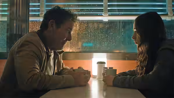 A man and woman sit across a diner table, talking over cups
