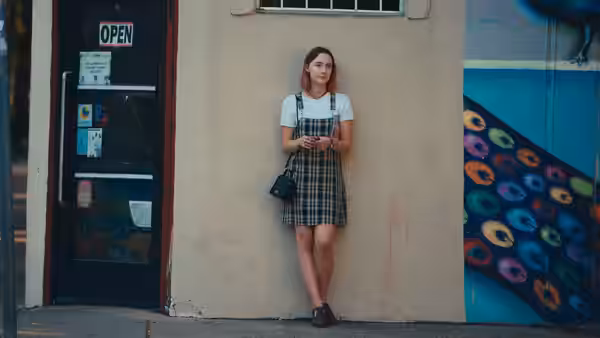 Lady Bird (Saoirse Ronan) leans against a wall outside a shop holding her phone