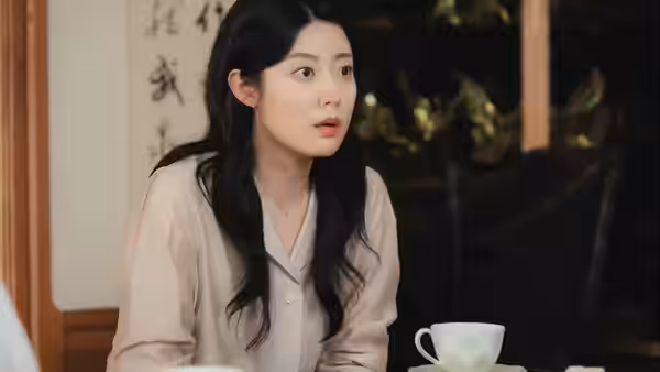 A woman in a pale blouse reacts across a table with a teacup
