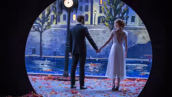 Man in a suit and woman in white dress hold hands on rose petals before painted cityscape
