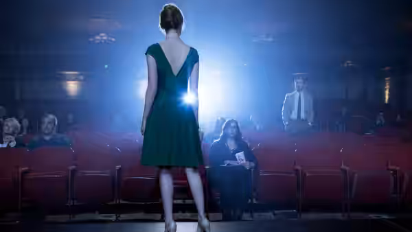 Woman in green dress stands in theater aisle facing a man under bright spotlight