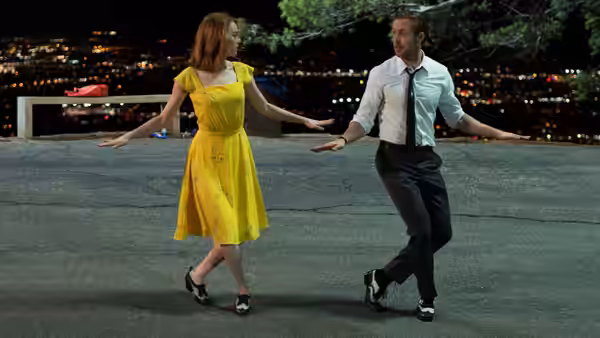 Man and woman in yellow dress dance with arms outstretched on rooftop at night