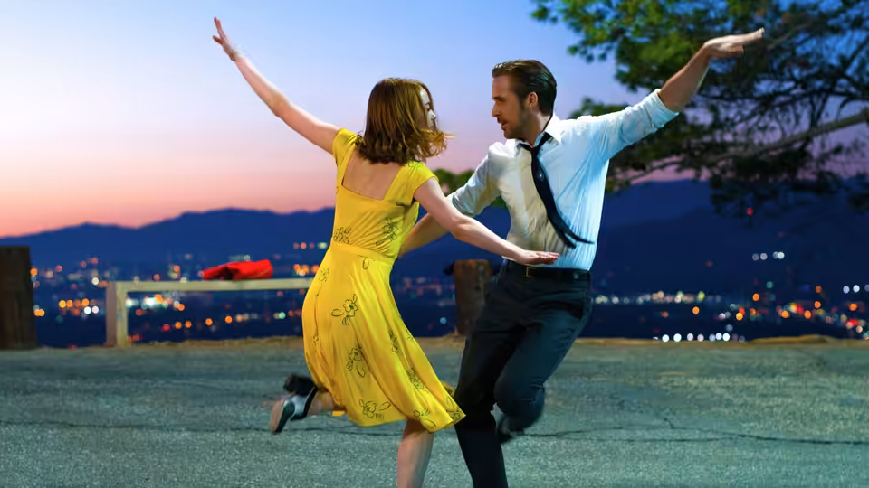 Man and woman in yellow dress dance with arms raised on outdoor platform at dusk