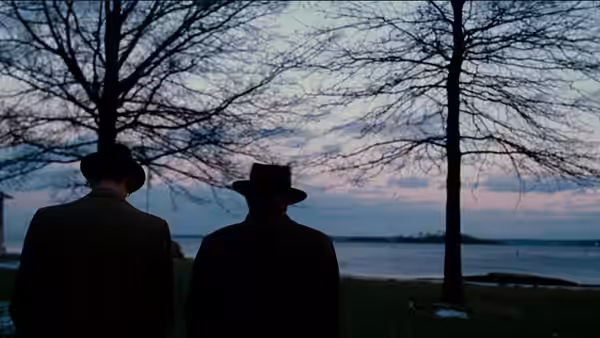 Two men in hats stand beneath bare trees beside a river at dusk