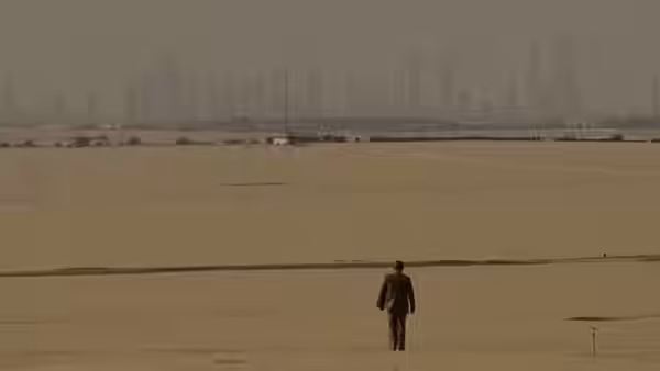 A lone man walks across a barren expanse toward a distant city skyline