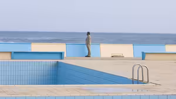 A lone man stands beside an empty swimming pool facing the sea