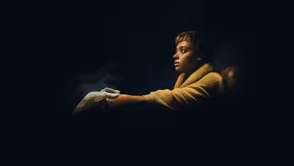 A woman in a yellow robe extends her bandaged hand in darkness