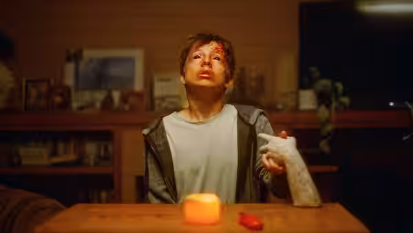 A bloodied young man with a bandaged hand sits at a wooden table