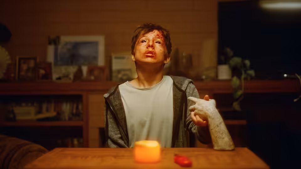 A bloodied young man with a bandaged hand sits at a wooden table