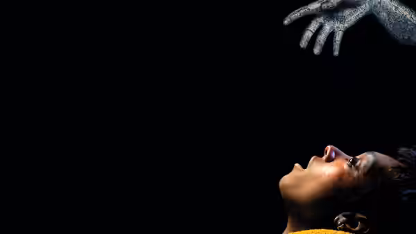 A woman with glitter on her face reaches upward against darkness