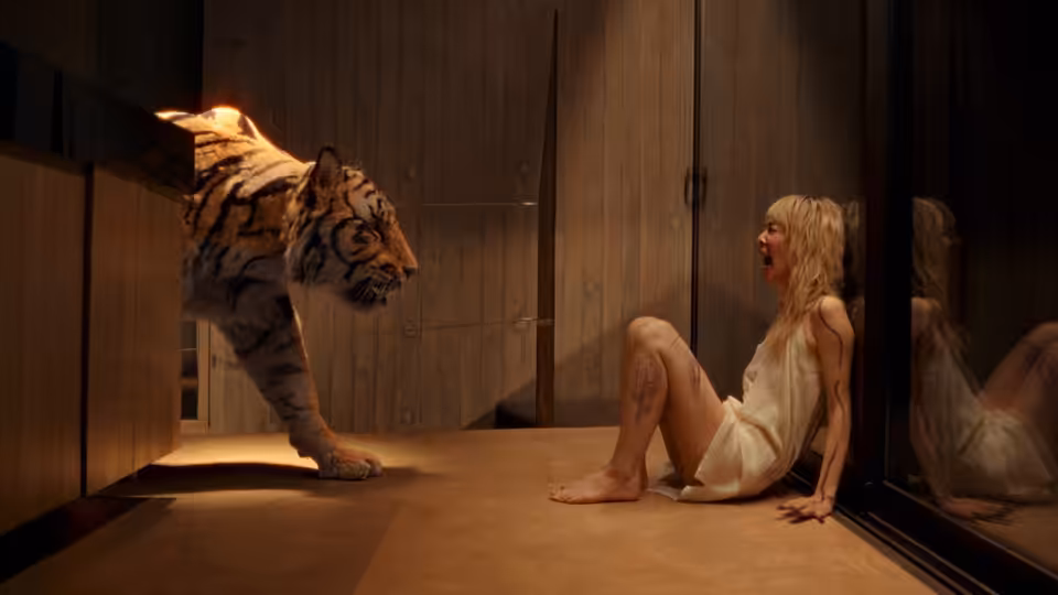A blonde woman sits against a wall as a tiger emerges from a cabinet