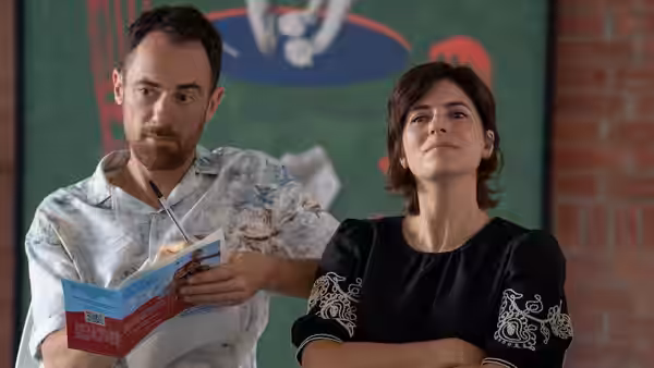 A man reads an open book beside a woman standing with folded arms