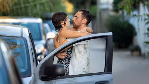 A man and woman embrace beside an open car door on a tree-lined street