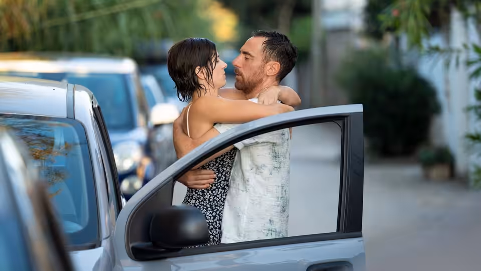 A man and woman embrace beside an open car door on a tree-lined street
