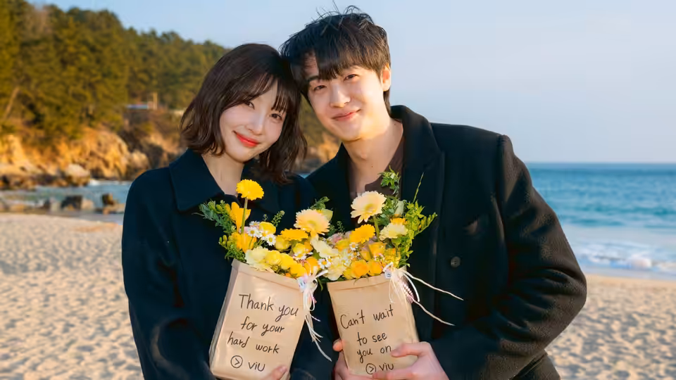 A young couple stand together on a beach, holding bouquets of yellow flowers