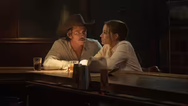 A cowboy-hatted man and a woman lean toward each other across a bar