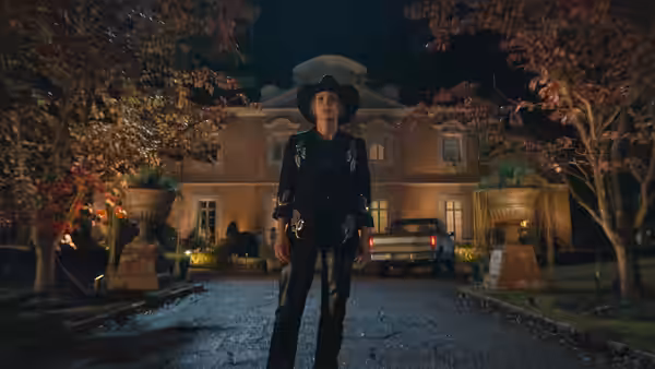 A cowboy-hatted figure stands in a driveway before a large house at night