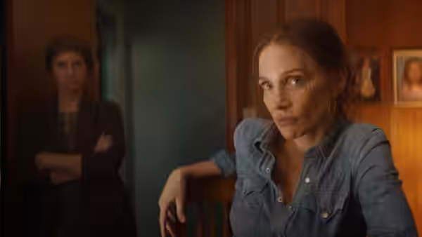 A woman in a denim shirt sits in the foreground while another watches from a doorway
