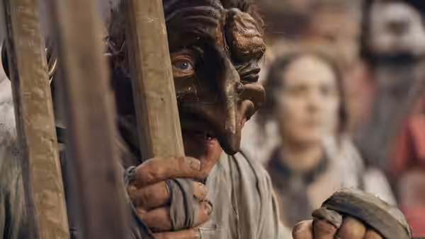 A man in a grotesque mask peers between wooden beams as a woman watches