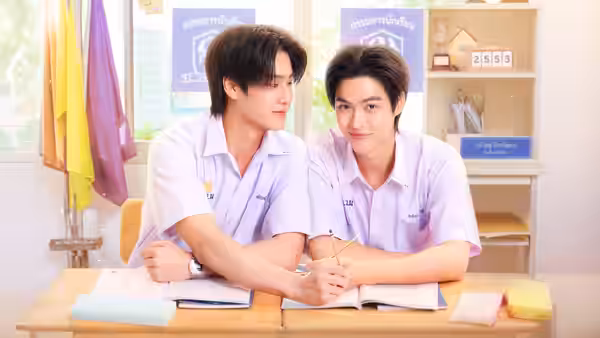 Two schoolboys share a notebook at a classroom desk, one writing as the other leans close