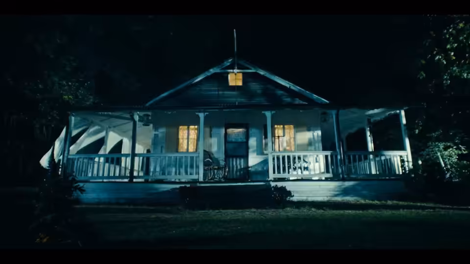 A wooden house with a lit porch stands among dark trees at night
