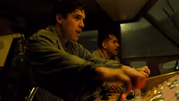 Two men operate a control console inside a dimly lit vehicle cabin