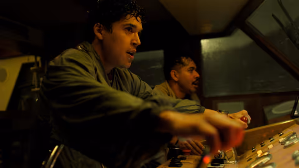 Two men operate a control console inside a dimly lit vehicle cabin