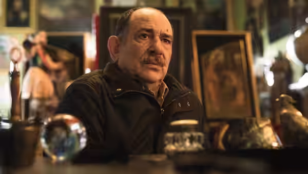 A mustached man sits at a counter among framed pictures and decorative objects