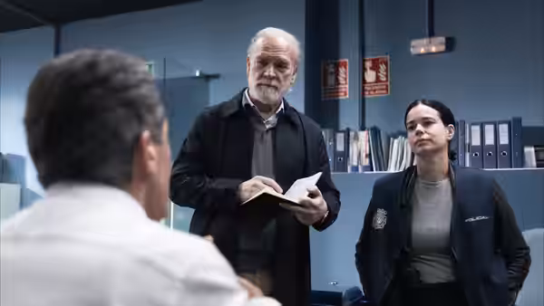 An older bearded man and a uniformed woman question a seated person in an office