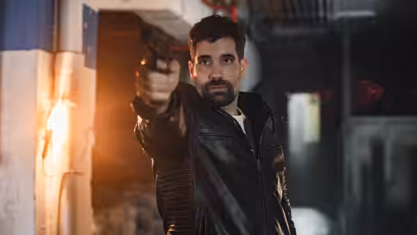 A bearded man in a leather jacket points a handgun down a corridor