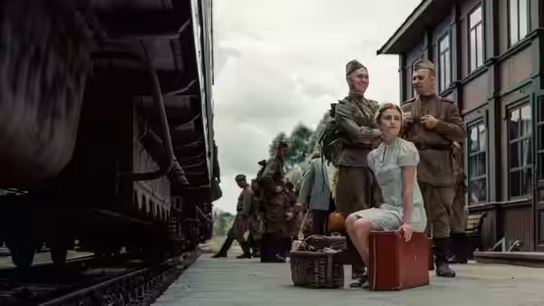 A young woman sits on a suitcase between two uniformed men on a train platform