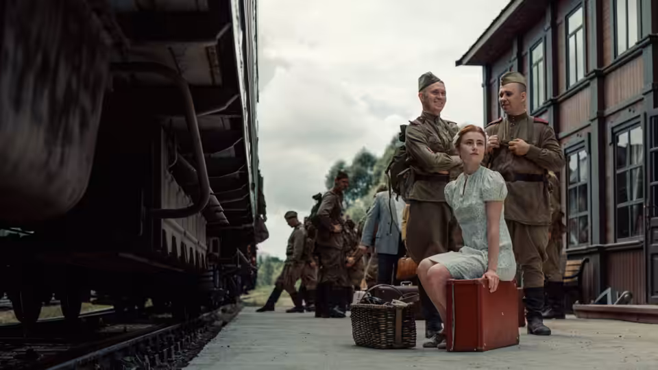 A young woman sits on a suitcase between two uniformed men on a train platform