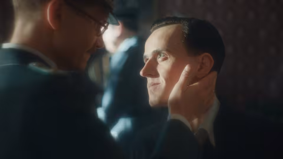 A man cups another man's face as they gaze at each other in a dim room