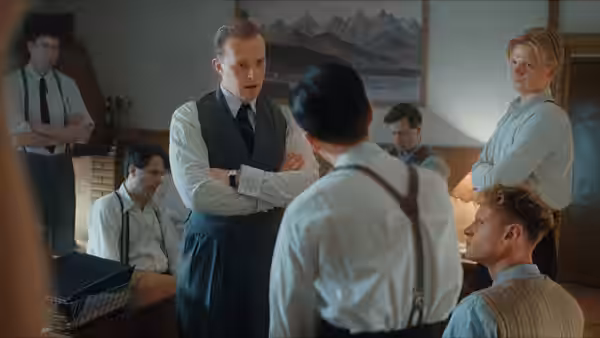 A man stands with folded arms before a group gathered around a table
