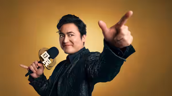 A man in black holds a microphone and points toward the camera against a gold backdrop