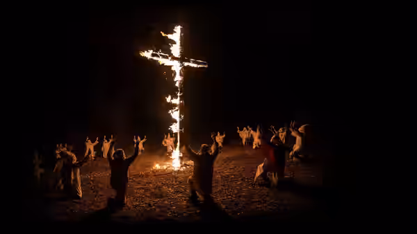 robed figures kneel with arms raised around a burning cross at night