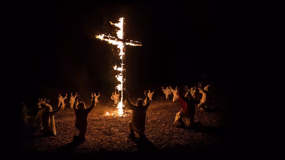 robed figures kneel with arms raised around a burning cross at night