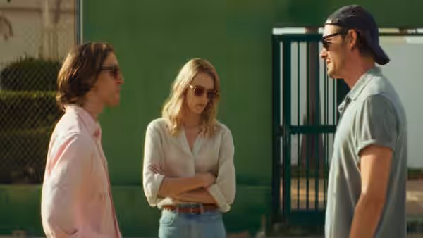 Anne (Stacy Martin) stands between two men outside a green building