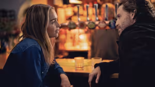 Ivy (Emilia Clarke) and Diego (Édgar Ramírez) face each other across a table in a warmly lit bar