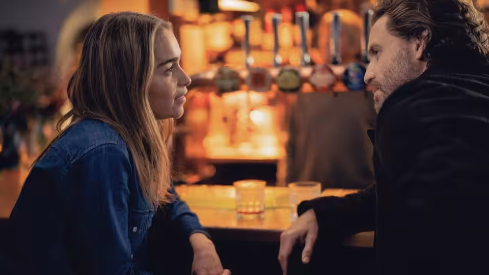 Ivy (Emilia Clarke) and Diego (Édgar Ramírez) face each other across a table in a warmly lit bar