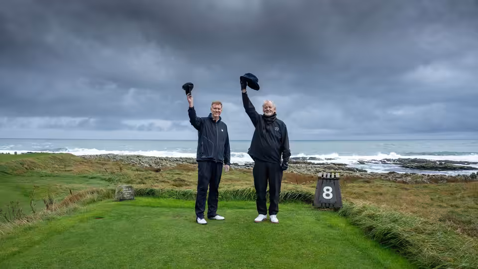 Two golfers raise their caps beside the eighth tee, with ocean waves under storm clouds