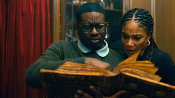 A man and a braided woman peer into an open book beside red curtains