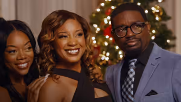A woman smiles between a friend and a bespectacled man beside a decorated Christmas tree