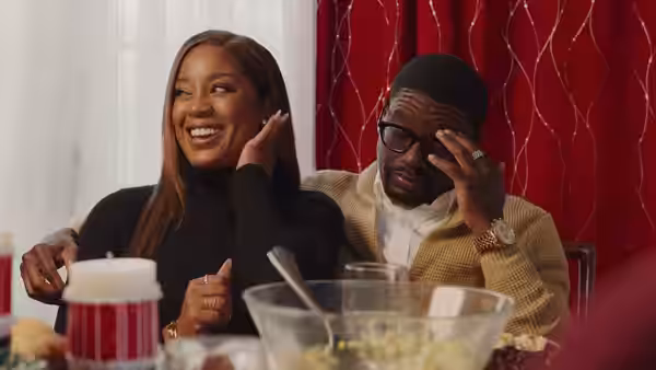 A woman laughs beside a bespectacled man at a dinner table as he rubs his eye